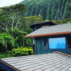 House with grey roof and green walls surrounded by lush trees and mountains.