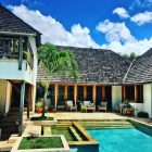 Luxurious villa with a swimming pool, palm tree, and lounge chairs under a bright blue sky.