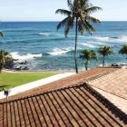 Oceanfront view with palm trees and a tiled roof overlooking blue waves and a sandy coastline.